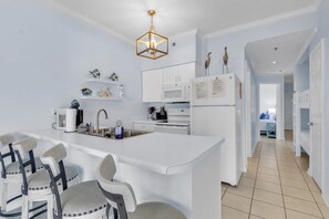 A modern kitchen with white cabinetry, a central island with bar stools, a hanging light fixture, and a view into the adjacent bedroom. - Tradewinds 1208 — Beach‑Front Escape with Sweeping 12th‑Floor Views (Orange Beach)