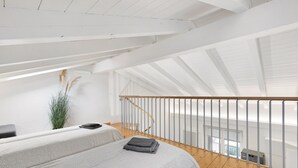Une pièce aux hauts plafonds, dotée d’un plancher en bois, d’un lit recouvert d’une couvre-lit à motifs gris, d’une petite plante et offrant une vue sur l’extérieur.