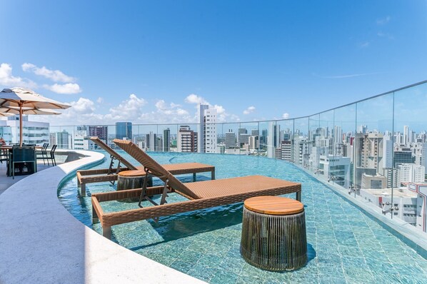Une piscine à débordement située sur le toit, avec des chaises longues et une table, offrant une vue panoramique sur la ville.