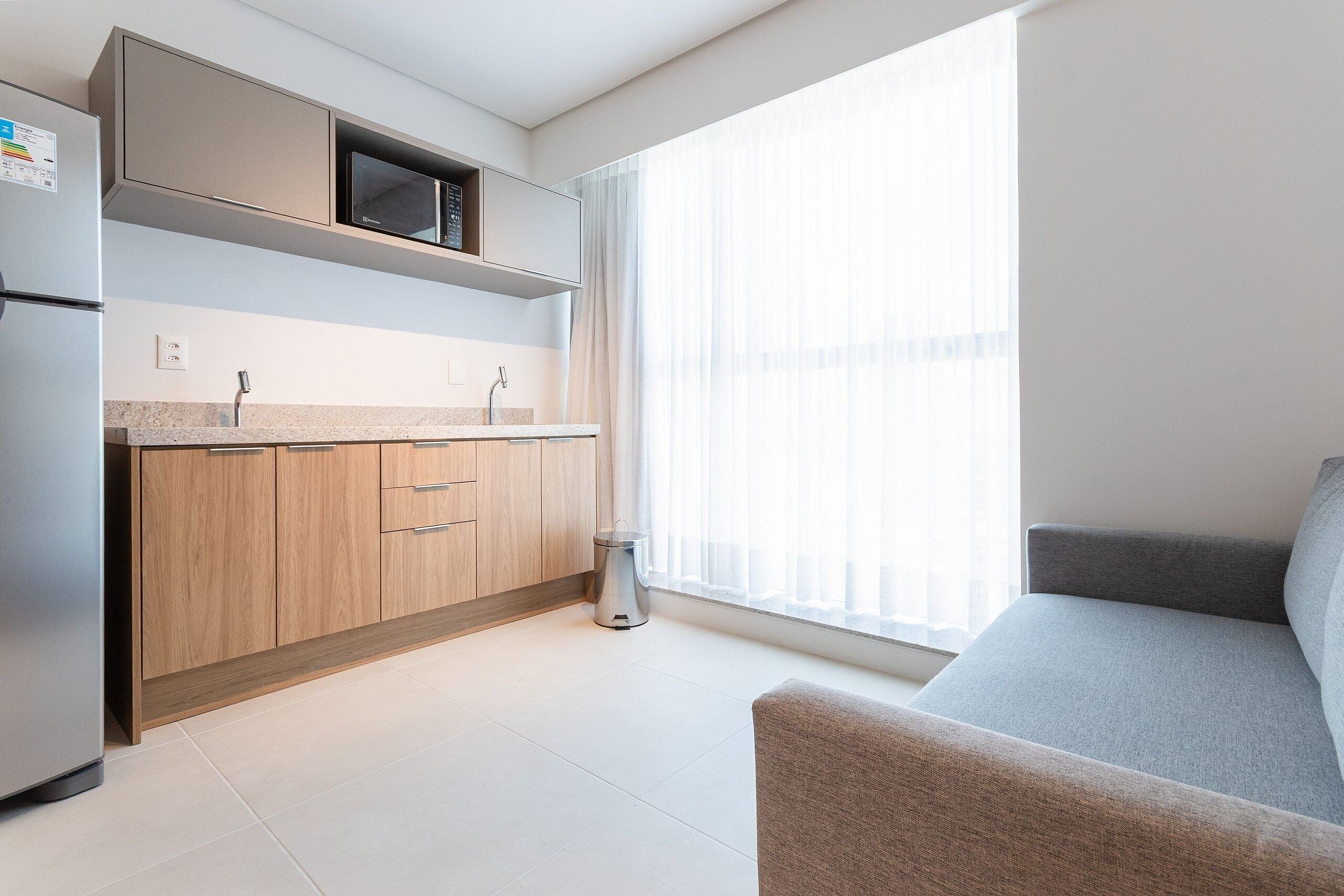 A modern kitchen with a built-in microwave, wooden cabinets, a stainless steel refrigerator, and a window with sheer curtains.