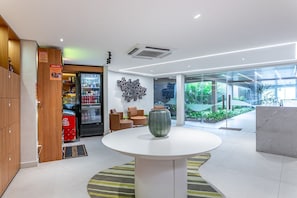 Un hall d’entrée moderne avec un distributeur automatique, une table ronde agrémentée d’un vase et un mur de verre donnant sur un espace verdoyant.