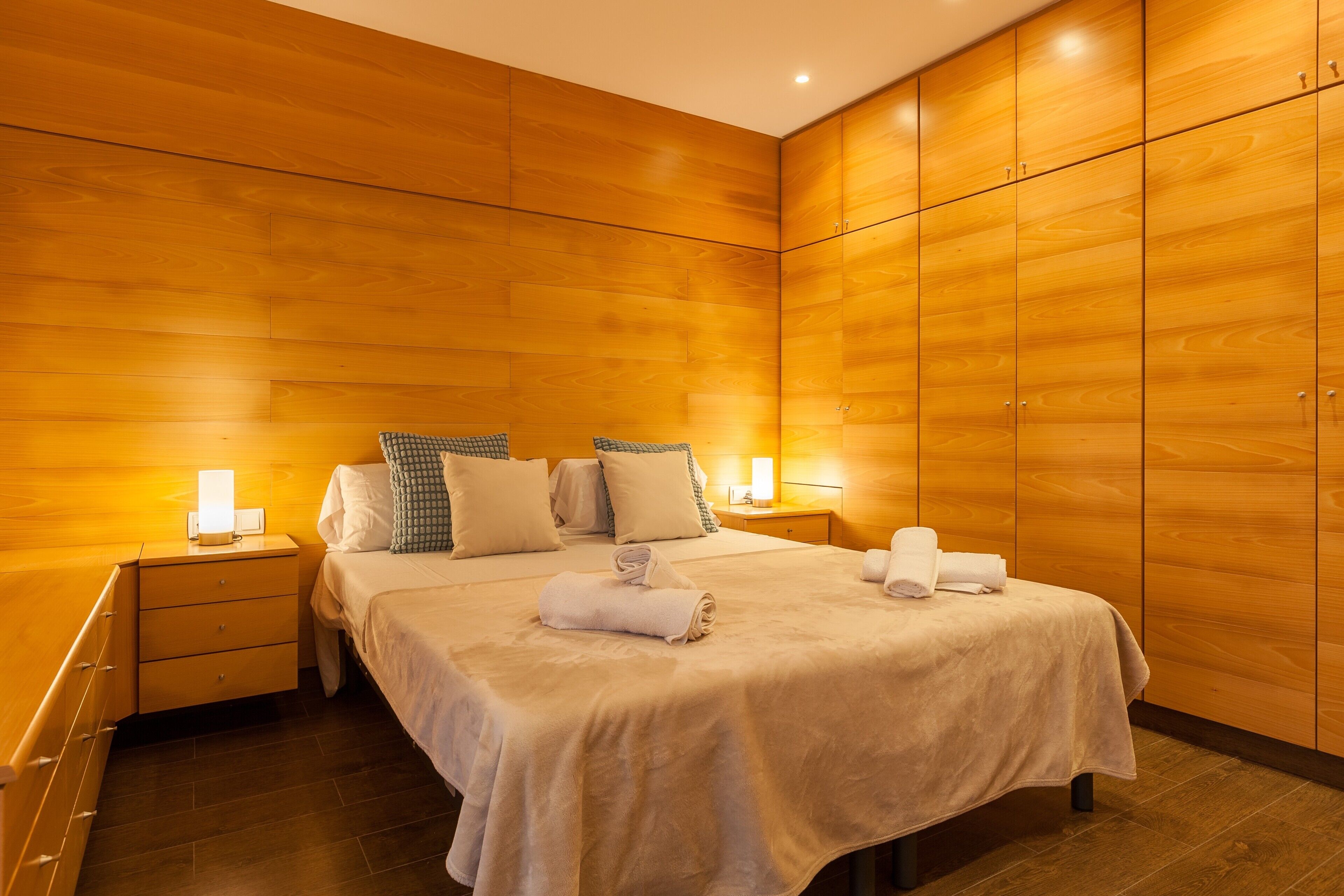 A hotel room with a wooden panel wall, a bed with white linens, two bedside tables with lamps, and a wooden wardrobe.