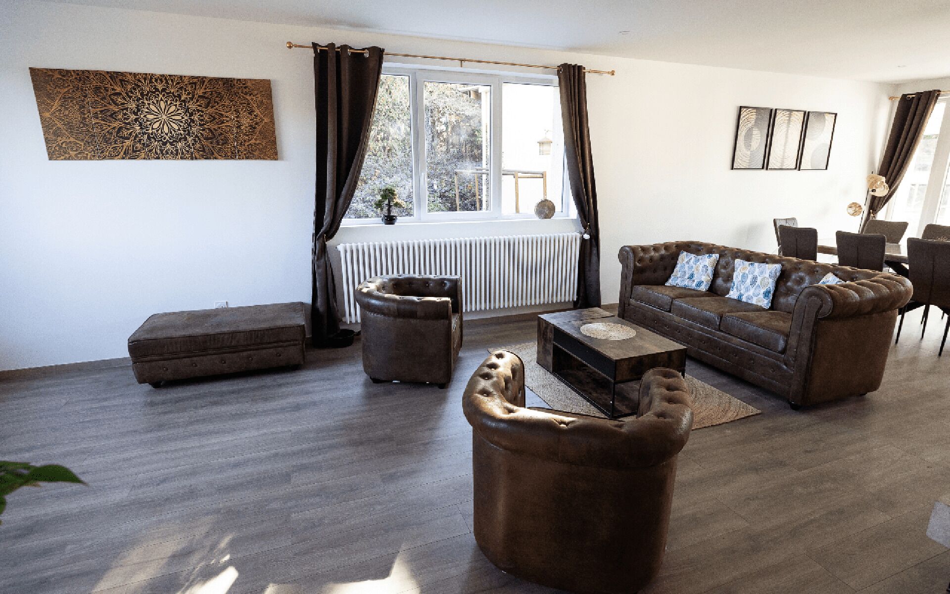 Een woonkamer met een bruine bank, een salontafel en een bruin voetenbankje. Er is een raam met gordijnen, een radiator en een plant op de vensterbank.