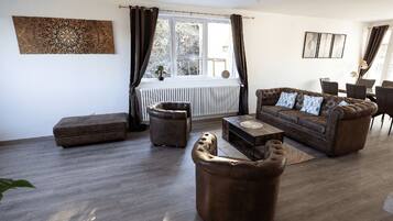 Een woonkamer met een bruine bank, een salontafel en een bruin voetenbankje. Er is een raam met gordijnen, een radiator en een plant op de vensterbank.