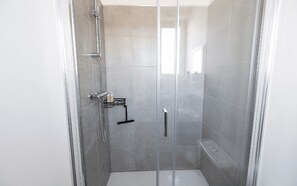 A glass-enclosed shower stall with a handheld showerhead, a towel rack, and a small shelf with toiletries.