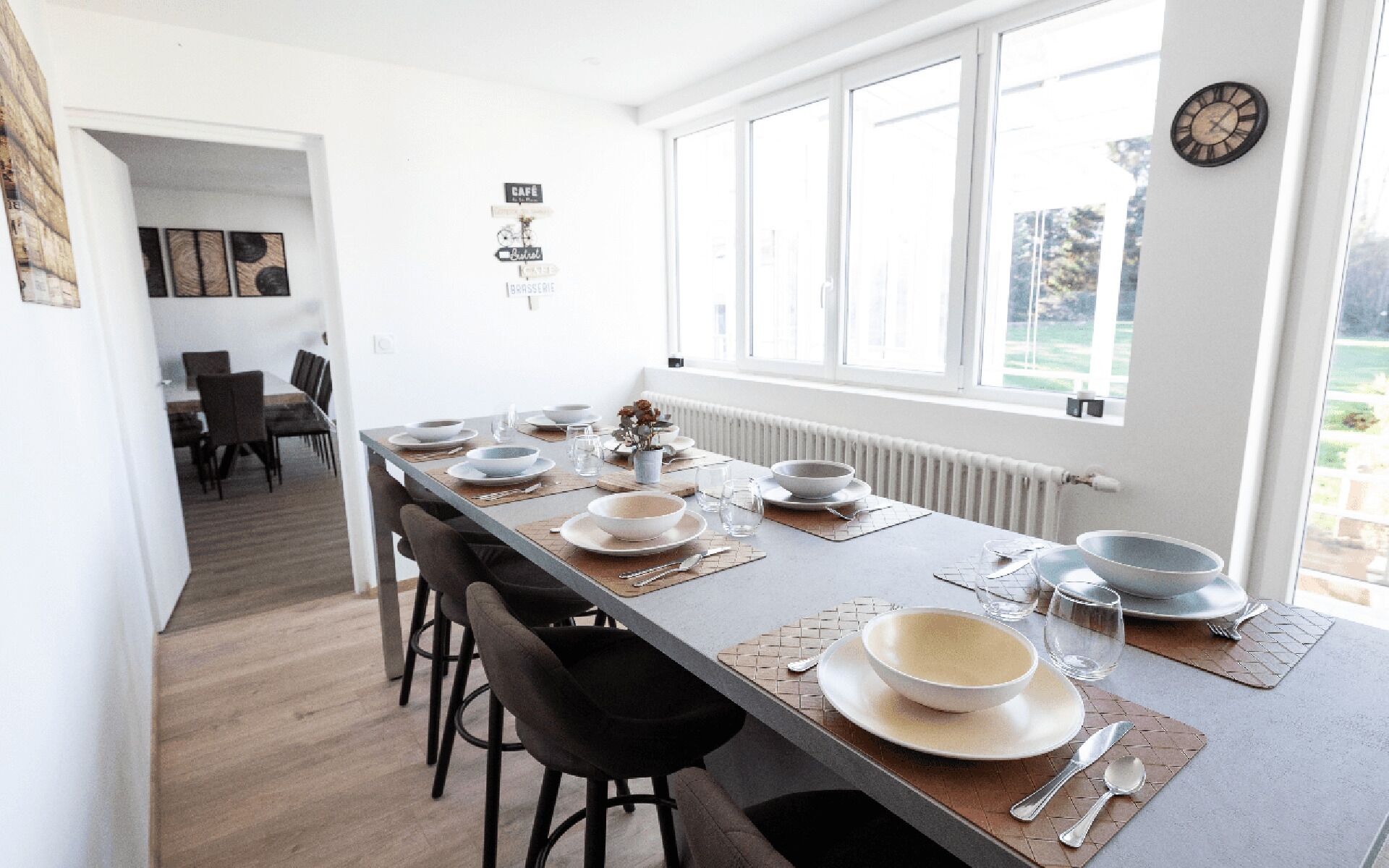A modern dining area with a long table set for six, a clock on the wall, and a view of the outdoors through large windows.