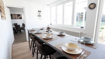 A modern dining area with a long table set for six, a clock on the wall, and a view of the outdoors through large windows.