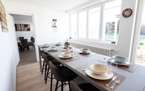 A modern dining area with a long table set for six, a clock on the wall, and a view of the outdoors through large windows.