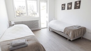 A bedroom with two single beds, a radiator, a window with a view of trees, and three wall-mounted pictures.