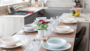 A modern kitchen with a long dining table set for a meal, featuring white and light blue bowls, clear glasses, and a potted plant with red flowers.