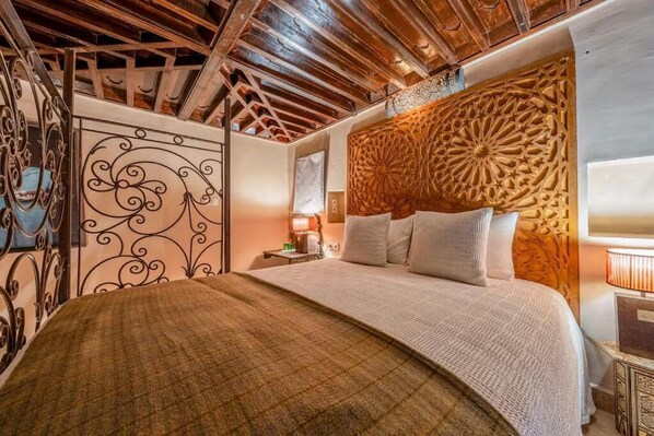 Une chambre à coucher avec un plafond en bois, une tête de lit décorative, un lit recouvert de linge de maison blanc et une table de chevet avec une lampe.