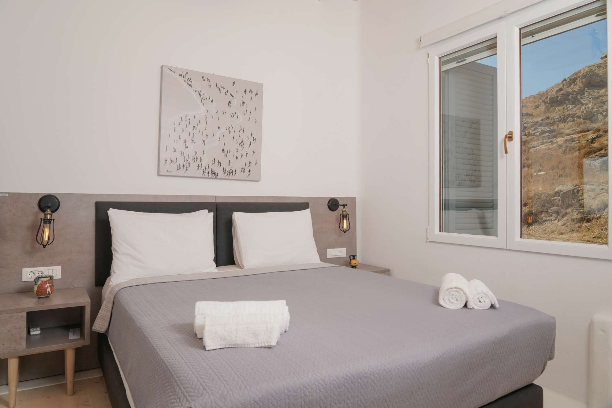 A bedroom with a bed, two bedside tables, a wall-mounted light fixture, a window with a view of rocky terrain, and a framed abstract artwork.