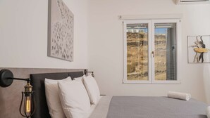 Et soveværelse med seng, et natbord, en væglampe, et vindue der viser et landskab, og et billede i ramme på væggen.