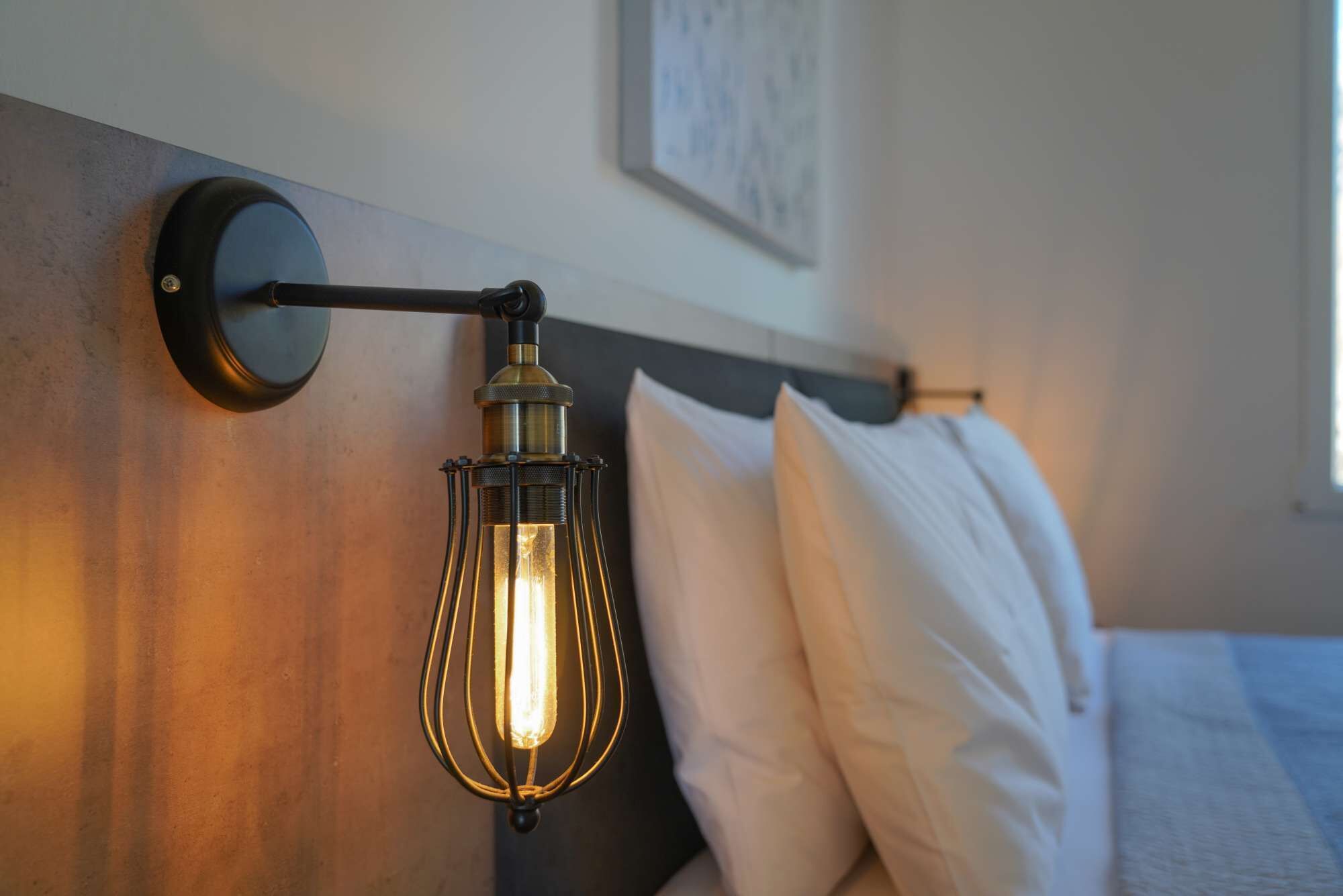 A wall-mounted light fixture with a visible bulb, a bed with white bedding, and a framed picture on the wall.