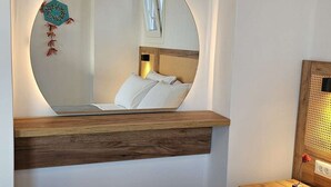 Una habitación de hotel con un espejo redondo, una cama con cubierta acolchada, una mesita de noche de madera y un taburete pequeño de mimbre.