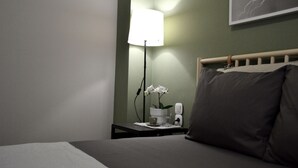 A neatly made bed with a pillow, a bedside table with a lamp, a small vase with flowers, and a framed picture on the wall.