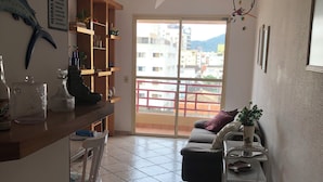 Un salon avec un canapé, une table basse et un balcon donnant sur des immeubles. On y trouve également un ventilateur de plafond et un panneau accroché au mur.