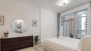 Une chambre moderne avec un grand lit, une commode, un miroir rond et une vue sur de hauts immeubles par la fenêtre.