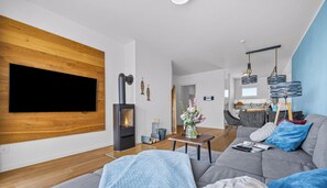 Ein modernes Wohnzimmer mit Kamin, Sofa, Fernseher und einem Essbereich, im Hintergrund ist eine Küche zu sehen.