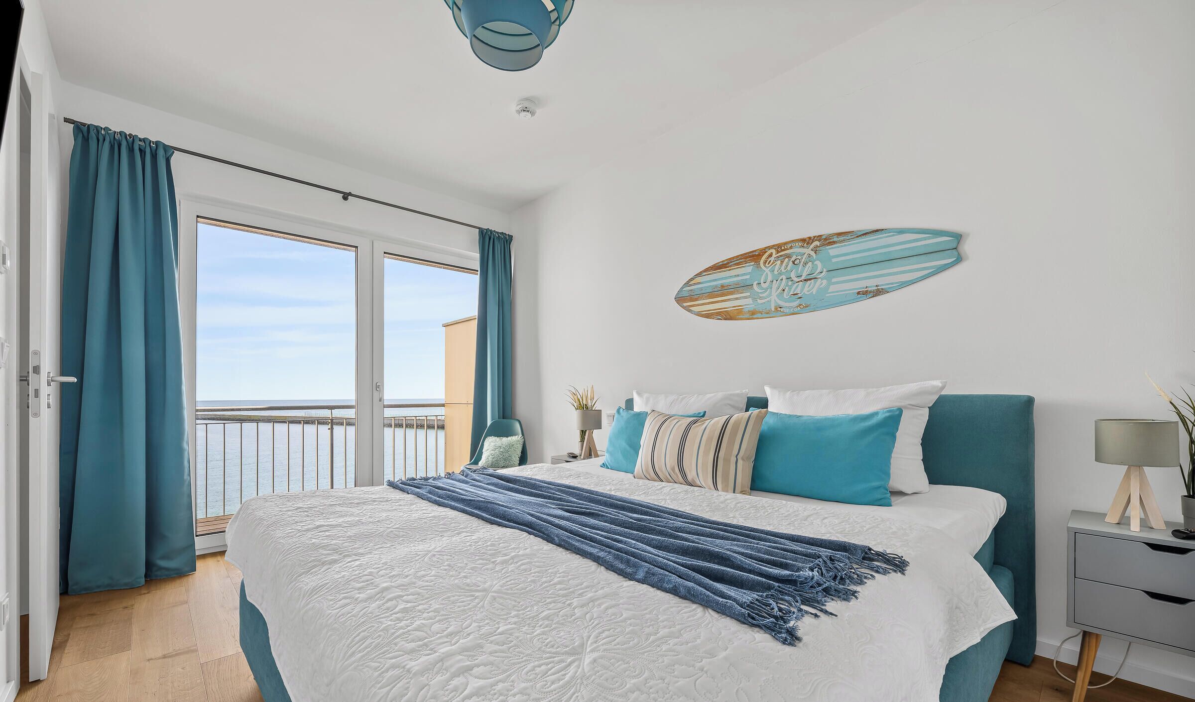 Een slaapkamer met een groot bed, uitzicht op zee door het raam, een surfboard decoratie en een nachtkastje met een lamp.