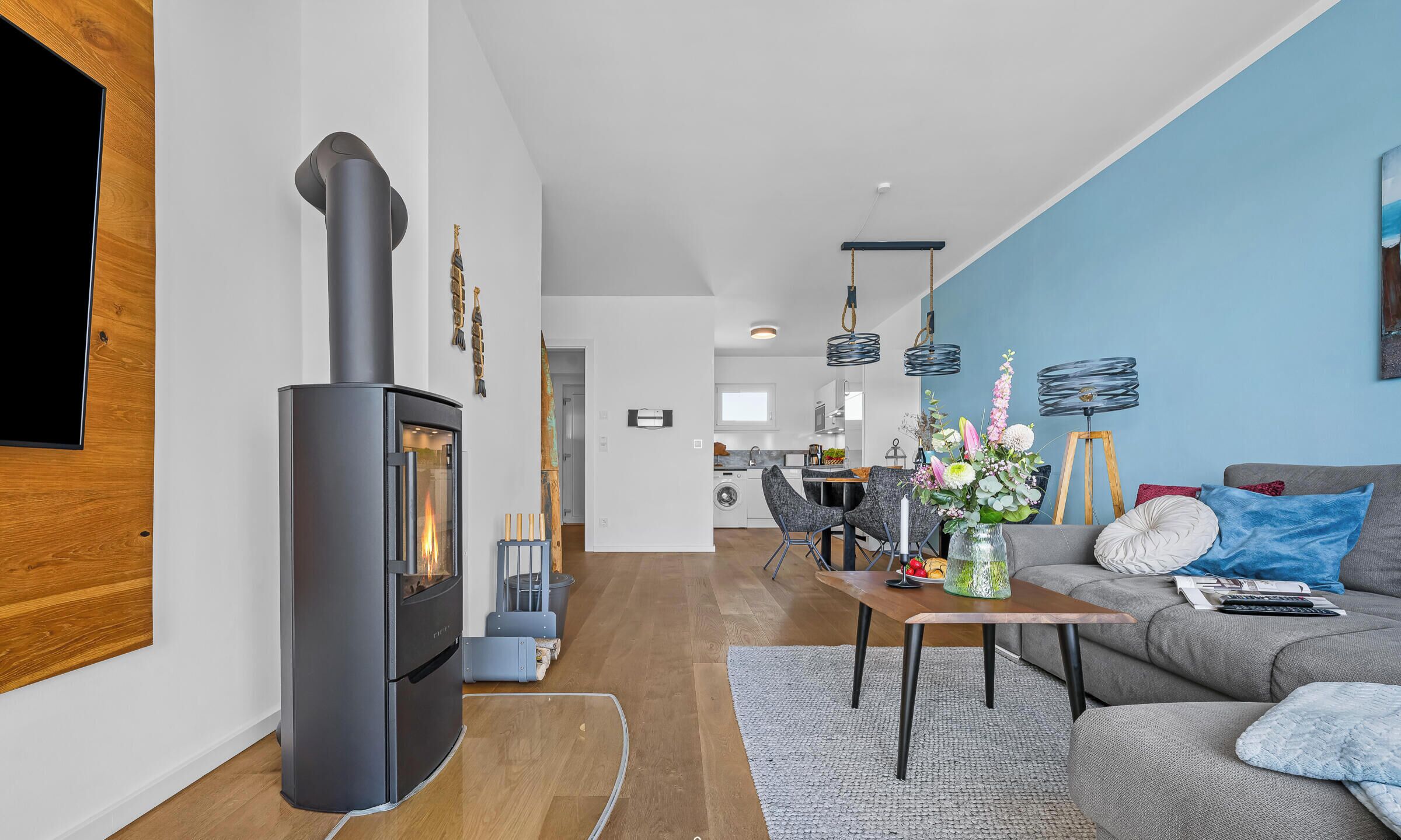 A modern living room with a wood-burning stove, a grey sofa, a dining area, and a kitchen in the background.