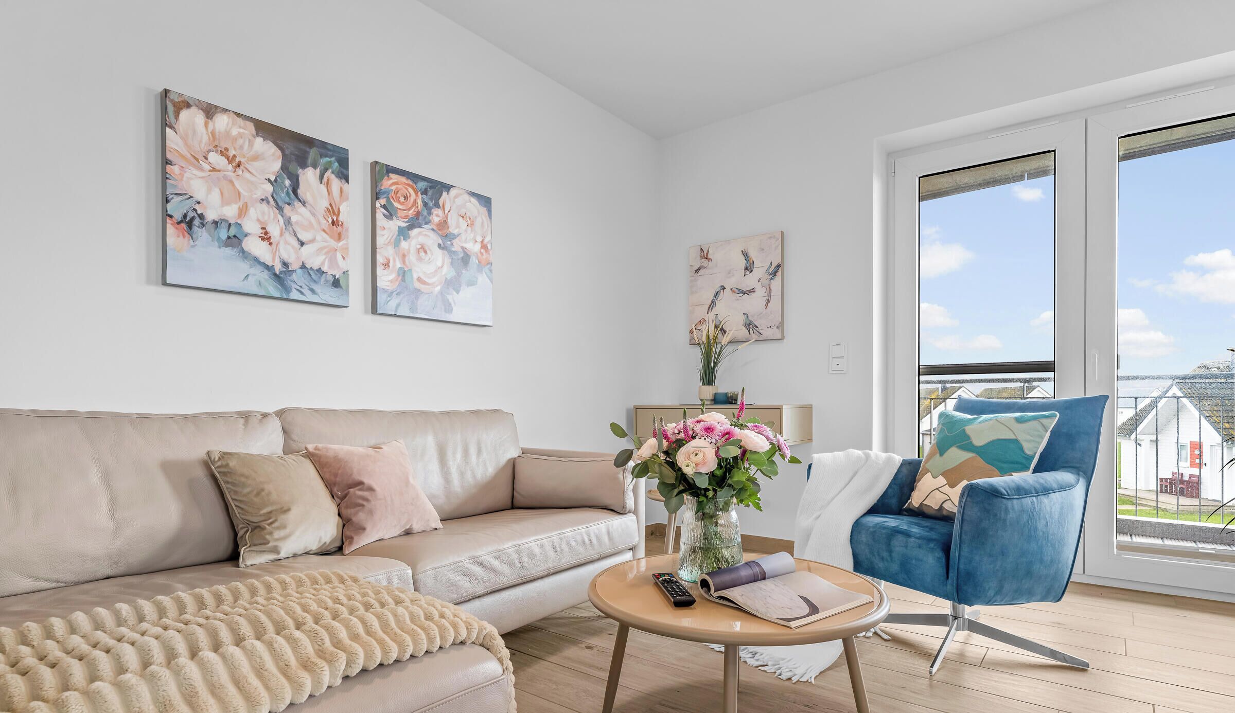Ein modernes Wohnzimmer mit einem beigefarbenen Sofa, einem blauen Sessel, einem runden Couchtisch und zwei Blumenbildern an der Wand.