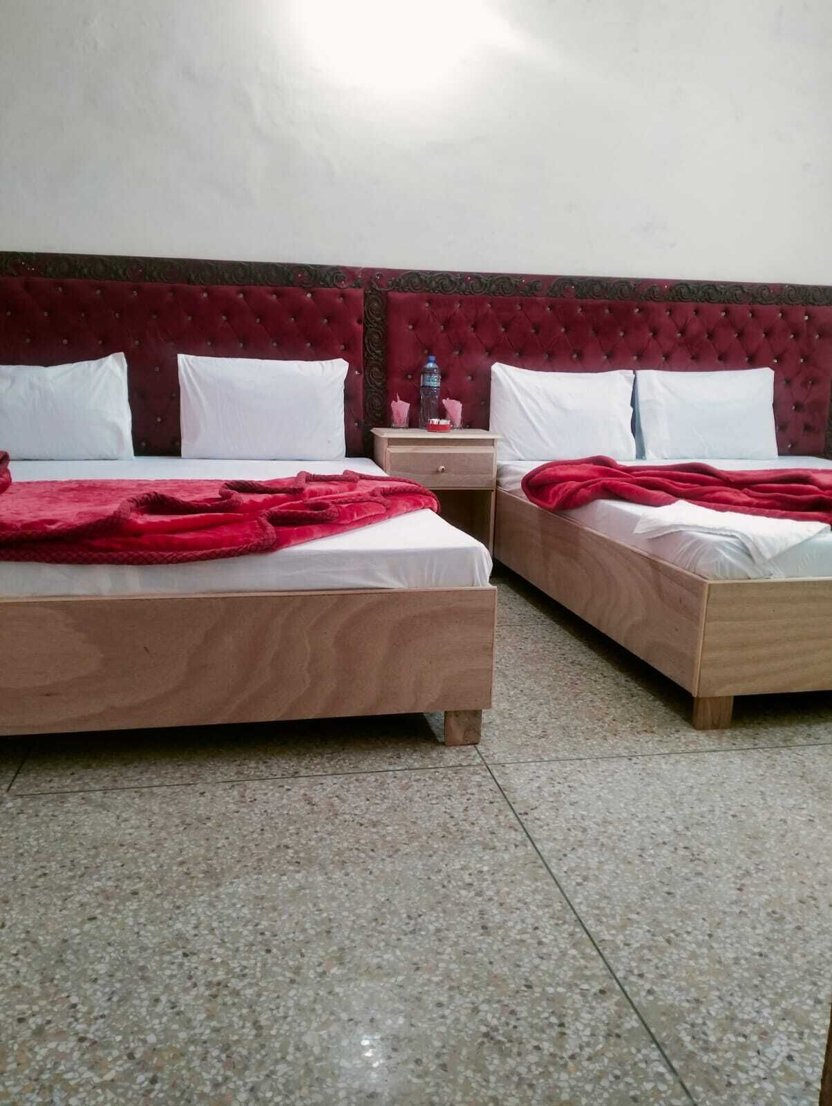A hotel room with two wooden beds, each with white linens and a red blanket. There is a nightstand with a water bottle and a tissue box.