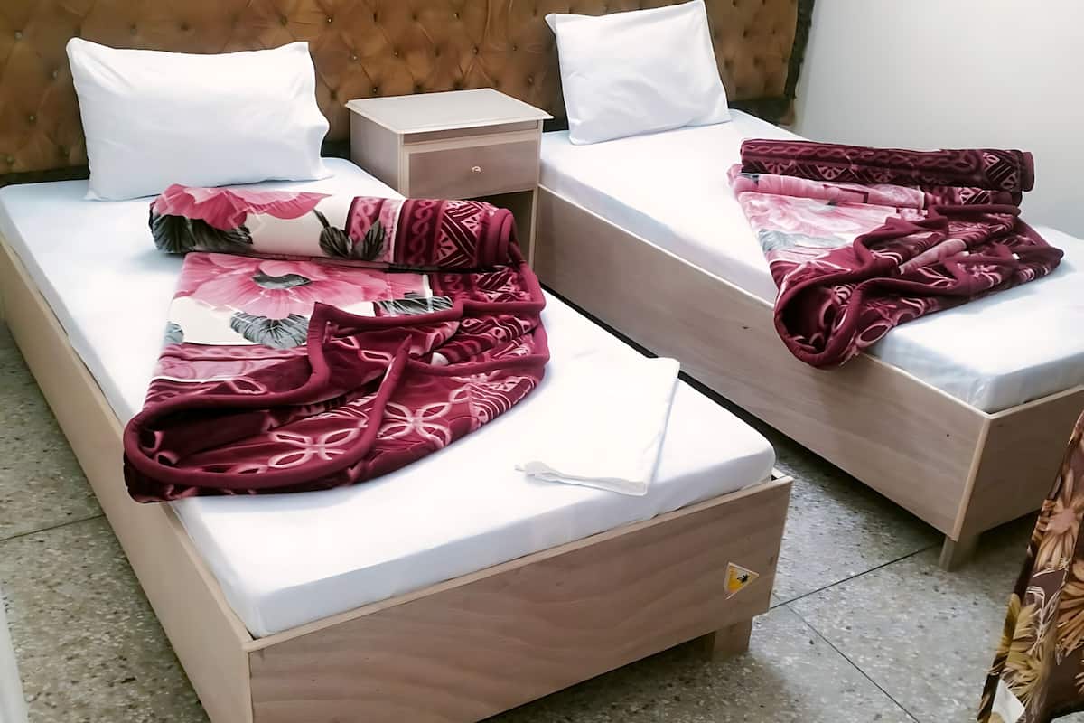 Quarto de hotel com duas camas, cada uma com colchão branco e cabeceira marrom. Duas almofadas brancas em cada cama, e um cobertor vermelho com padrão floral está dobrado sobre as camas.