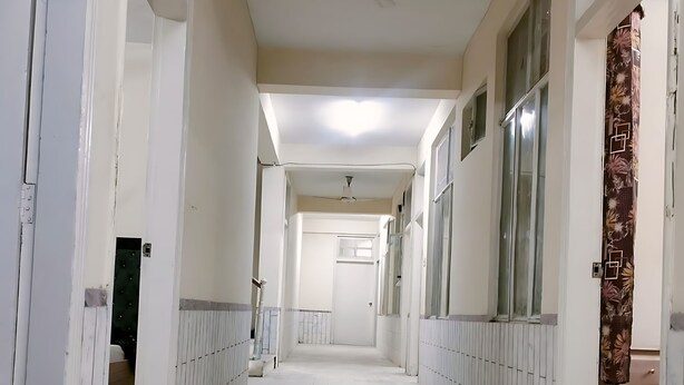 A long, narrow hallway with white walls and a marble floor. There are doors on both sides, and a ceiling fan is visible in the center. - Hotel The Prime (Rawalpindi)