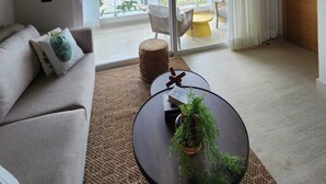 Un ampio soggiorno con soffitto in legno, una grande porta scorrevole in vetro che dà su un balcone, un divano con cuscini, un tavolino rotondo con una pianta e una poltrona gialla.