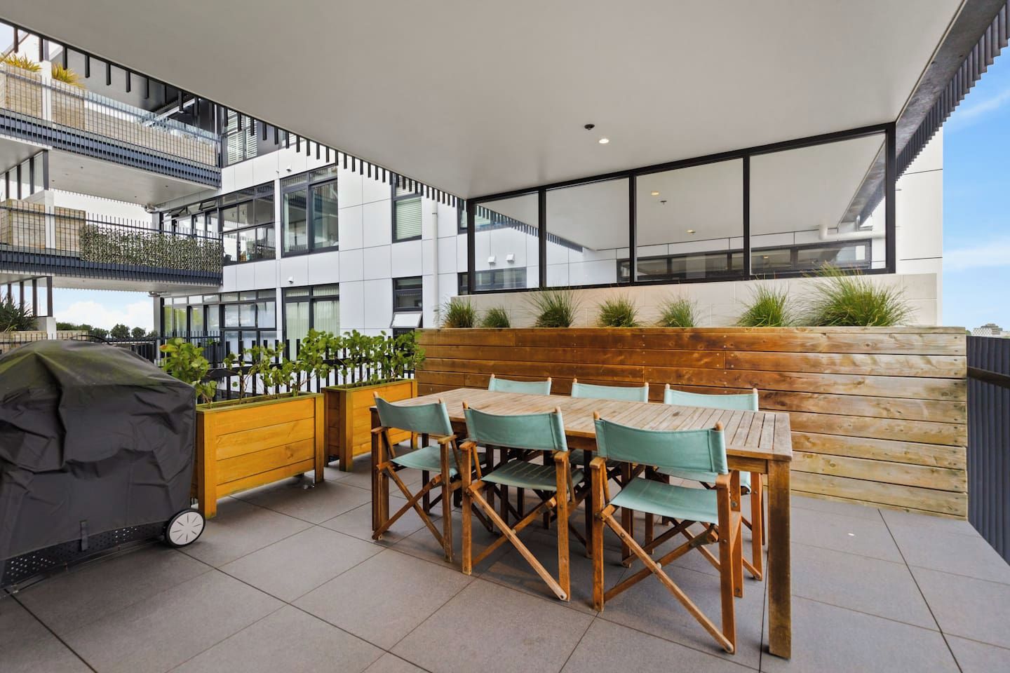 An outdoor dining area with a wooden table and chairs, a grill, and a view of a modern building with balconies.