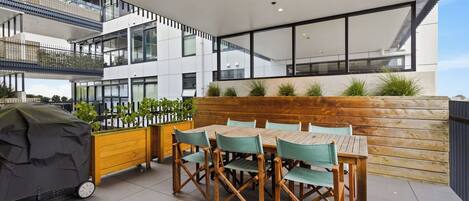An outdoor dining area with a wooden table and chairs, a grill, and a view of a modern building with balconies.