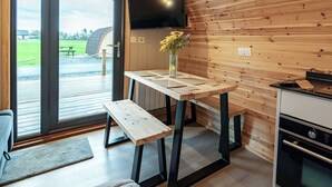 A modern kitchen with a wooden interior, a dining table with benches, a TV mounted on the wall, and a view of a green outdoor area. - 1 bedroom accommodation in Millom (Millom)