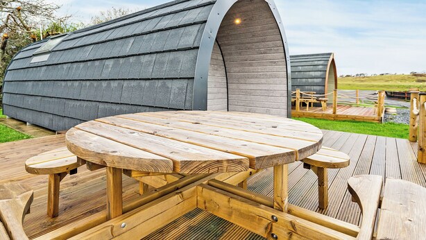 A wooden deck with a round table and benches, a covered structure in the background, and a grassy field. - 1 bedroom accommodation in Millom (Millom)