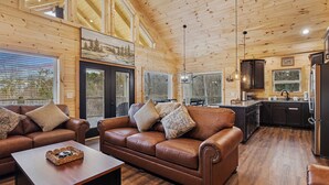A spacious living room with a brown leather sofa, a wooden coffee table, and a large window with blinds. - Rivers Retreat | Luxury New Cabin Near Gatlinburg (Sevierville)