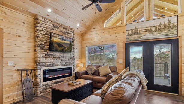 A cozy log cabin interior with a stone fireplace, leather sofa, and a large window offering a view of the outdoors. - Rivers Retreat | Luxury New Cabin Near Gatlinburg (Sevierville)