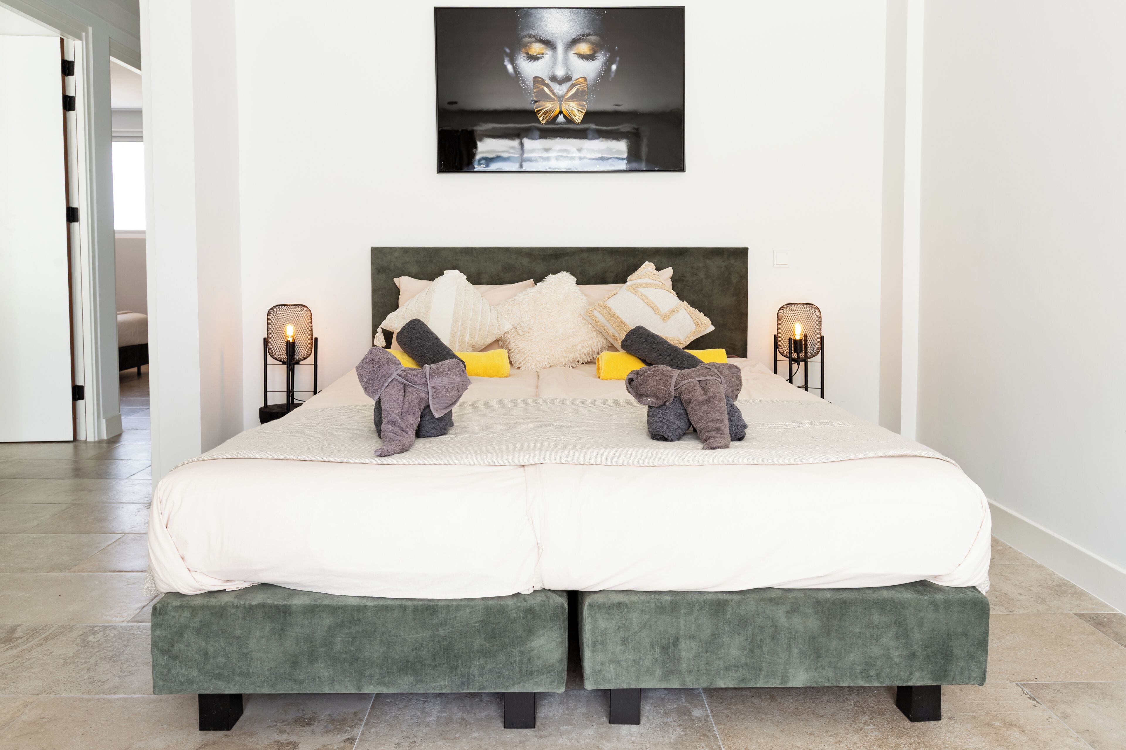 Una cama doble con cabecera tapizada de verde, dos mesitas de noche con lámparas y una obra de arte enmarcada sobre la cama.