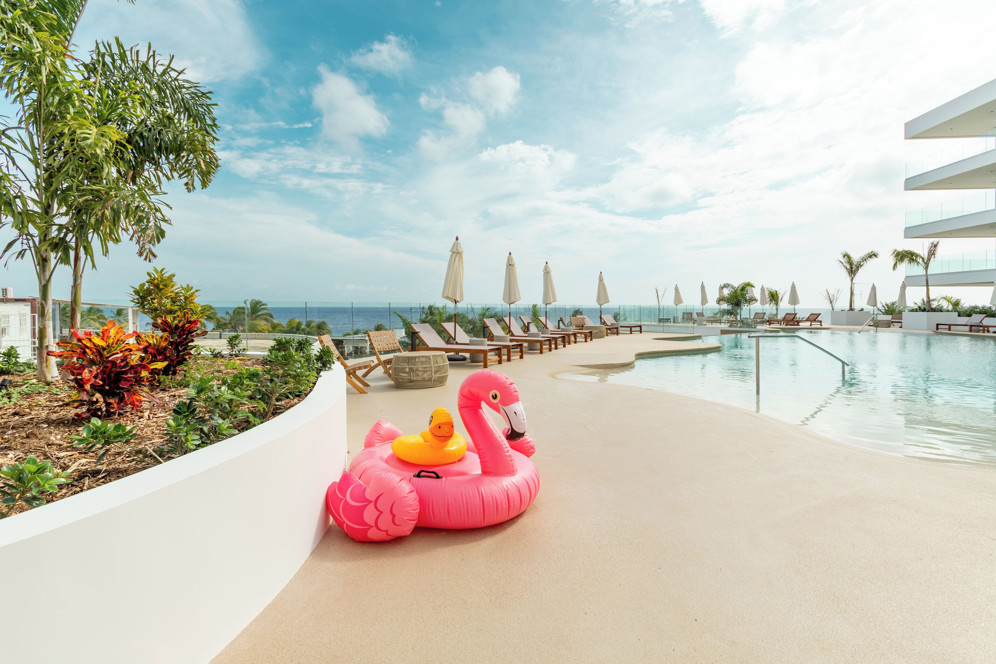 Zona de piscina con sillas de descanso, sombrillas y un flotador con forma de flamenco rosa.