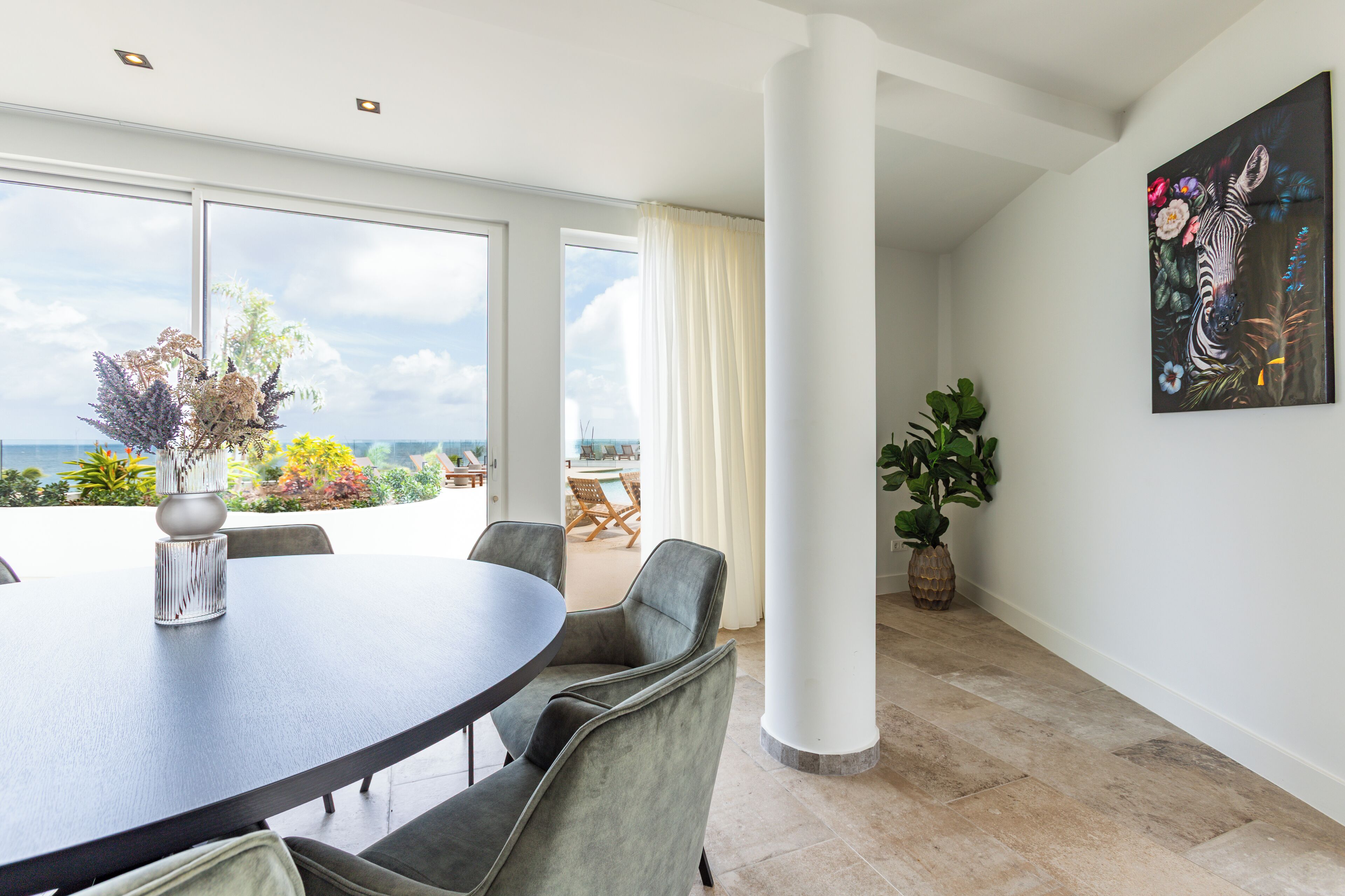 A dining area with a round table and chairs, large windows with a view, and a potted plant.