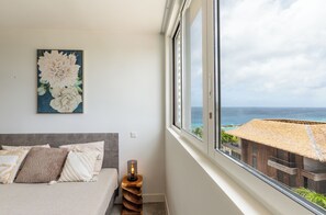A bedroom with a bed, pillows, a nightstand, a lamp, and a large window with a view of the ocean and a building with a thatched roof. - Mambo Beach Holiday Apartments (Willemstad)