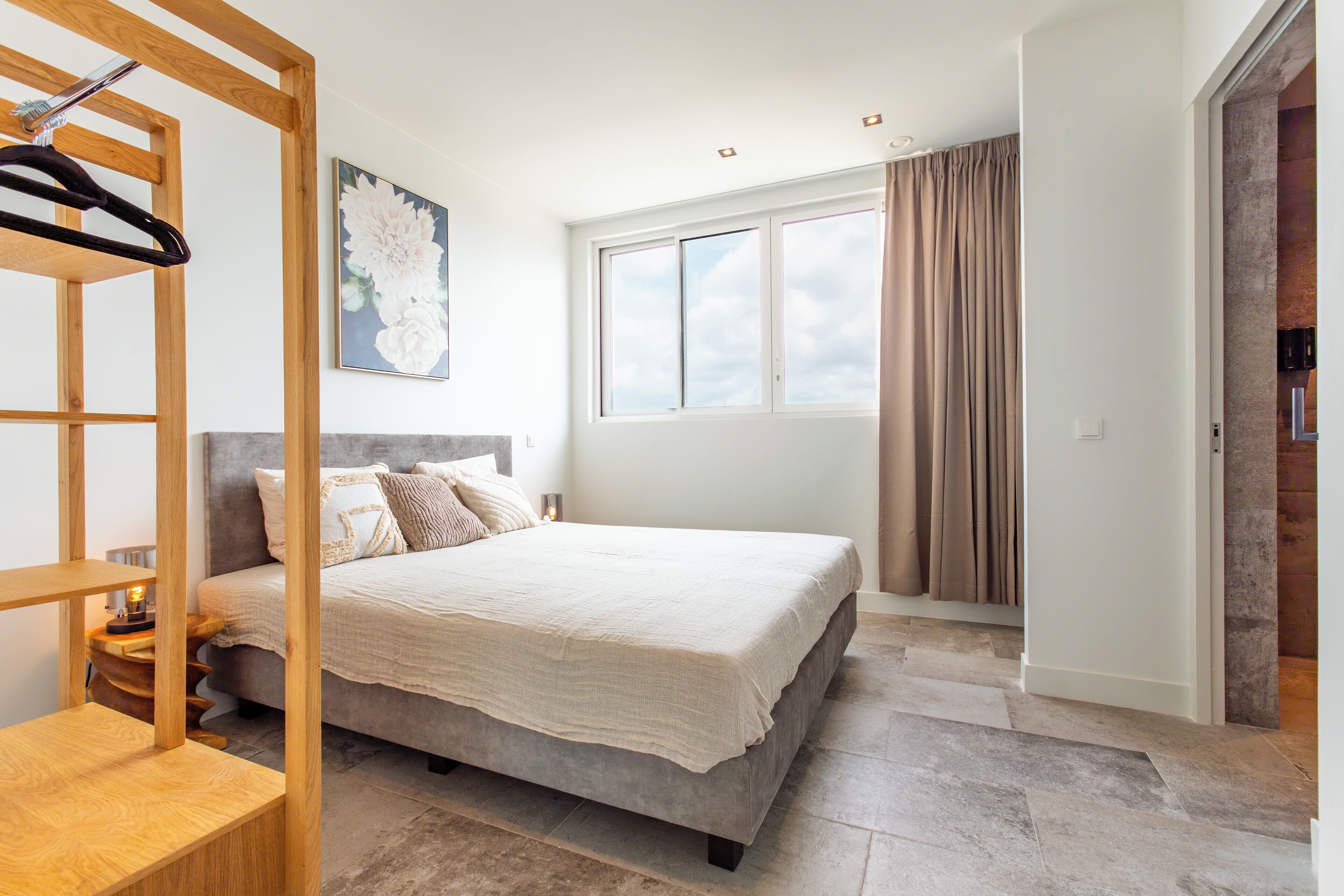 Un dormitorio con una cama, un estante de madera, un cuadro, una ventana y una puerta.