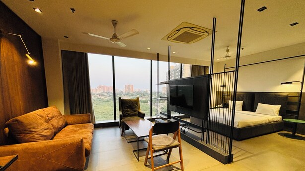 A modern hotel room with a large TV, a sofa, a chair, a bed, and a view of the city.