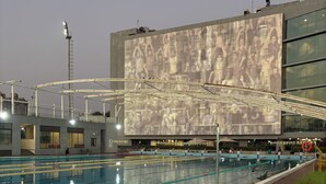 A swimming pool with clear lane dividers, a modern building with a large digital screen, and a wooden deck area with potted plants.