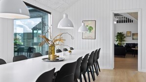 Un espace de salle à manger moderne avec une longue table blanche, des chaises noires et des luminaires suspendus. On peut voir un vase avec des branches et un bol doré sur la table.