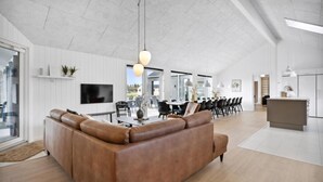 Un salon moderne avec un canapé en cuir marron, un coin repas avec des chaises noires et une cuisine avec des meubles blancs.