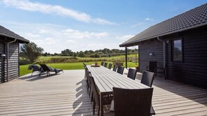 Eine Holzdielen-Terrasse mit langem Tisch und Stühlen, umgeben von einer Holzhütte mit einem Ziegeldach.