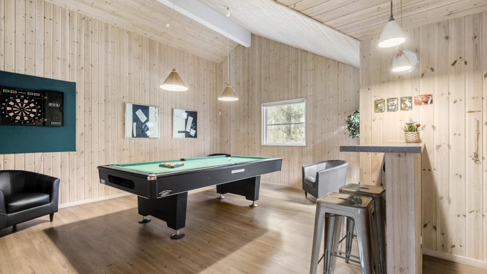 Ett biljardbord i ett rum med träpanel på väggarna och i taket, en darttavla och en bardisk med barstolar.