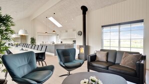 Un salon moderne comprenant un canapé, deux fauteuils, une table basse et un coin repas avec une longue table et des chaises. On aperçoit une cuisine à l’arrière-plan, avec une cuisinière et des placards.
