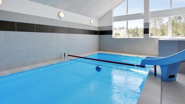 An indoor swimming pool with a blue water slide, tiled walls, and large windows.