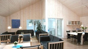Ein geräumiges Zimmer mit Holzwänden und -decke, eine große Schiebeglasdoor zum Balkon, eine Essecke mit Tisch und Stühlen sowie eine Sitzecke mit Sofas und Sesseln.